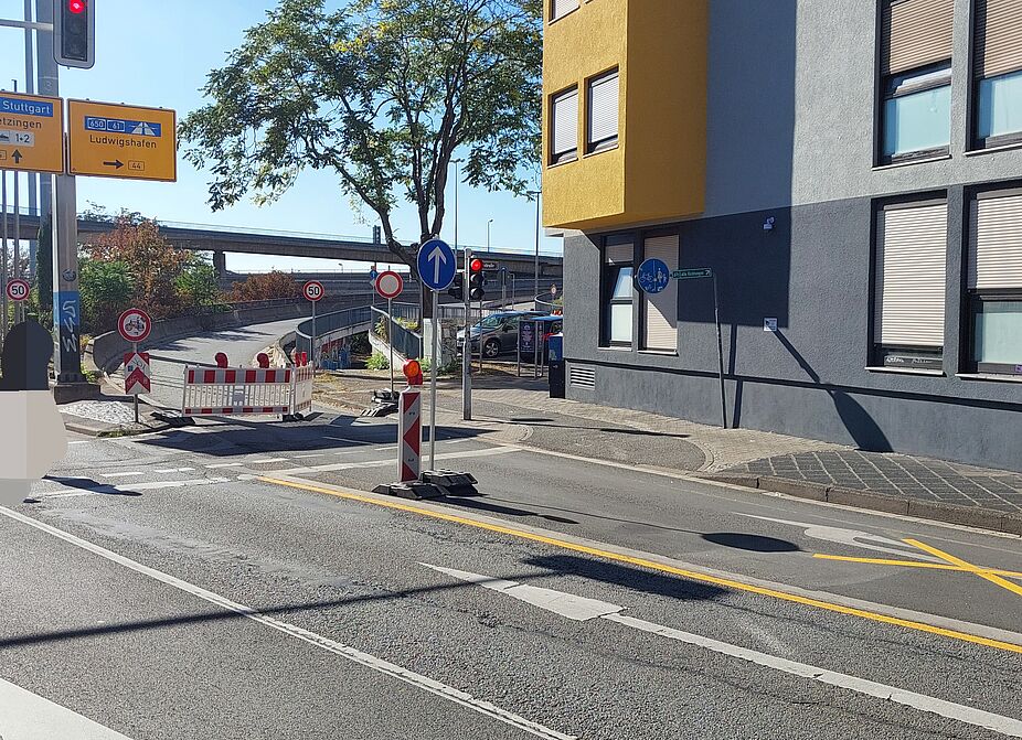 Auffahrt Richtung Ludwigshafen: Absperrungen statt klarer Wegweisung Kreuzung am Luisenring in Mannheim. Gelbe Wegweiser zeigen nach Stuttgart und nach rechts Richtung Ludwigshafen (B 44). Dahinter führt eine geschwungene Rampe nach oben, davor stehen ein rot-weißes Absperrgitter, Warnbaken und Verkehrsschilder. Rechts an einer grauen Hauswand hängt ein blaues Schild für einen gemeinsamen Geh- und Radweg. Auf der Fahrbahn sind gelbe Baustellenlinien und ein gelbes Kreuz markiert. Im Hintergrund verläuft die Hochstraße.