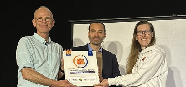 Nora Müller (links) vom ADFC SH übergibt die Plakette "Fahrradfreundliche Schule" an Mathias Kadow-Leder (Schulleiter Holstenschule) und Christian Wagner (Fahrradbeauftrager Holstenschule Neumünster)