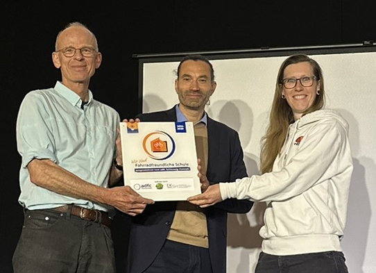 Übergabe Plakette "Fahrradfreundliche Schule" Nora Müller (links) vom ADFC SH übergibt die Plakette "Fahrradfreundliche Schule" an Mathias Kadow-Leder (Schulleiter Holstenschule) und Christian Wagner (Fahrradbeauftrager Holstenschule Neumünster)