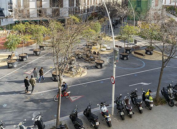 Straßenszene in einem Superblock in Barcelona. Ein Fahrrad fährt auf einer ehemaligen Straße. Es sind Bäume in Töpfen zu sehen. Ein paar Fußgänger gehen über die ehemalige Straße, die jetzt ein Radweg ist. Es ist ein Tempolimitschild zu sehen mit einer 10 darauf zu sehen, also 10 km/h. 