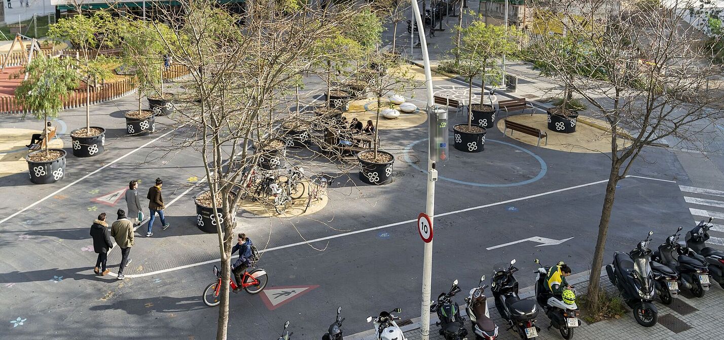 Straßenszene in einem Superblock in Barcelona. Ein Fahrrad fährt auf einer ehemaligen Straße. Es sind Bäume in Töpfen zu sehen. Ein paar Fußgänger gehen über die ehemalige Straße, die jetzt ein Radweg ist. Es ist ein Tempolimitschild zu sehen mit einer 10 darauf zu sehen, also 10 km/h. 