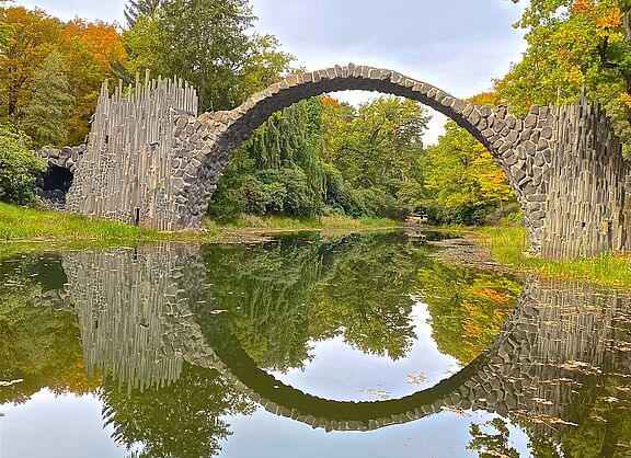 ein Foto der Rakotzbrücke, die sich halbkreisförmig über eine kleinen See erstreckt und sich darin spiegelt.