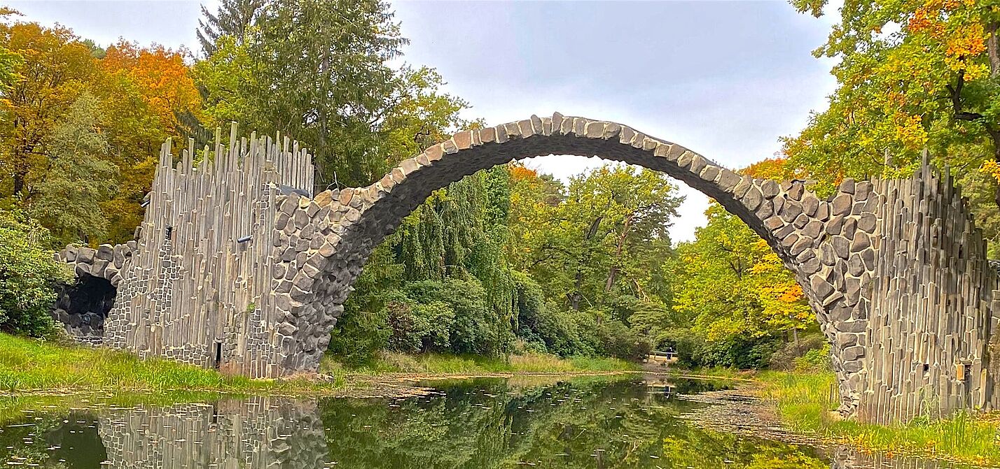 ein Foto der Rakotzbrücke, die sich halbkreisförmig über eine kleinen See erstreckt und sich darin spiegelt.