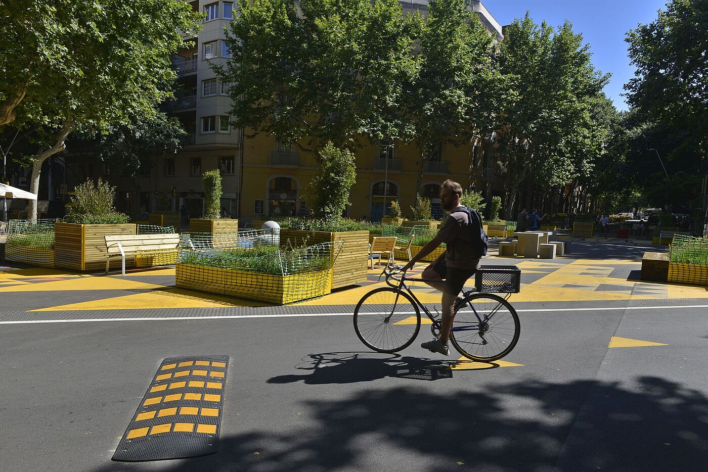 Ein Radfahrer, der durch den Superblock Poblenou fährt. Auf dem Boden ist gelbe Farbe aufgebracht, die Sonne scheint und auf der ehemaligen Straße bzw. Kreuzung stehen Hochbeete. Es ist eine entspannte Atmosphäre. 