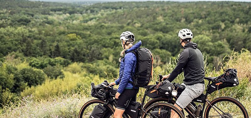 Bikepacking Eine Radfahrerin und ein Radfahrer stehen auf einer Anhöhe un blicken auf einen Wald.