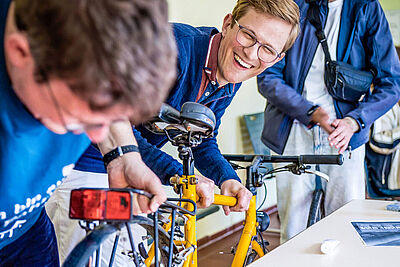 Menschen reparieren gemeinsam ein gelbes Fahrrad und lachen.